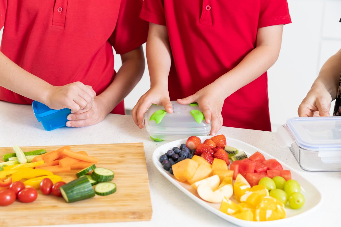 Children in school uniforms put chopped fresh fruit and vegetables into their lunchboxes