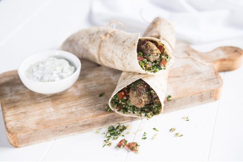 Image of two beef kofta wraps layered on top of a wooden cutting board with tzatziki on the side