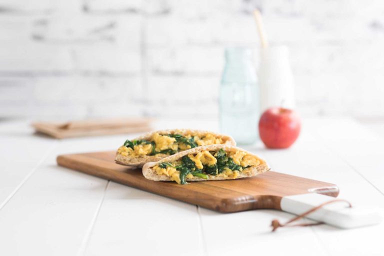 Two open breakfast pitas served on a wooden serving board with an apple, glass bottle of milk, glass bottle of water and brown paper napkins in the background.