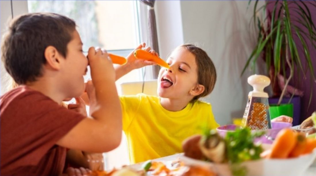 Two happy children playing with their carrots sit in front of a table with a grater and a wide range of vegetables