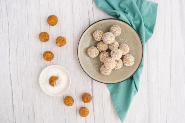 A shallow round bowl containing coconut coated bliss balls. The bowl is sitting on a blue napkin. To the left is 8 bliss balls not coated in coconut with 1 bliss ball sitting in a small bowl of coconut.