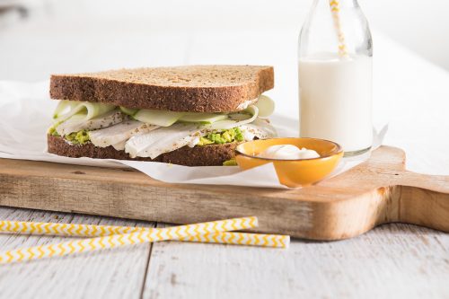Image of chicken avocado and cucumber sandwich served on a wooden cutting board with small yellow dish of aoli and a bottle of milk with a straw on the side