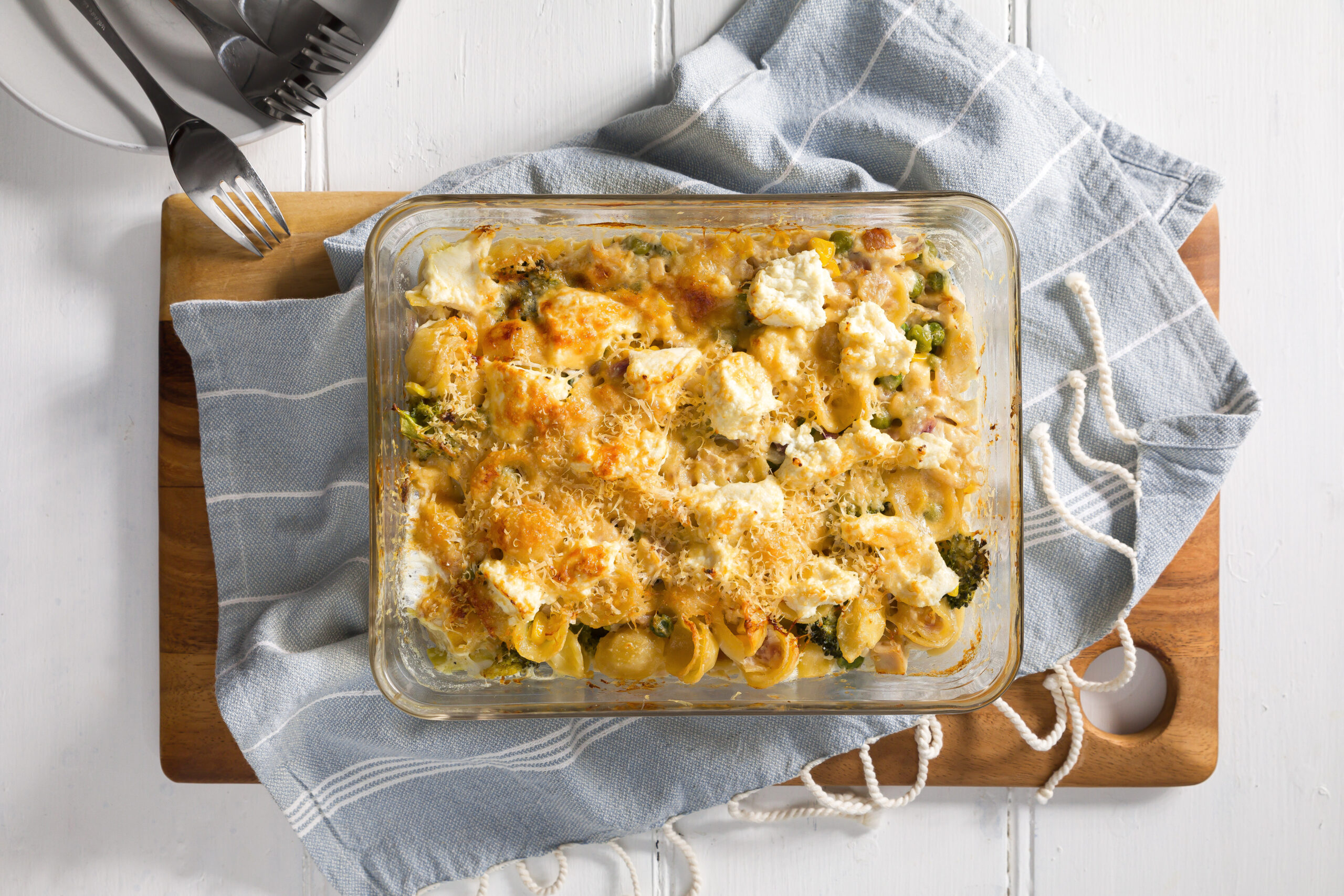 Chicken pasta bake in an glass oven-proof dish sitting on a blue napkin on a timber board with forks for eating on the left.    