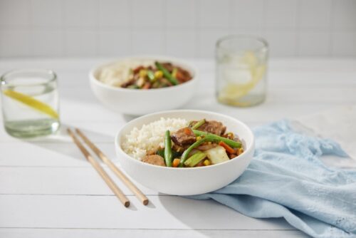 Two bowls of chicken and vegetables stir fry with rice on a table with chopsticks and glasses of water with lemon slices