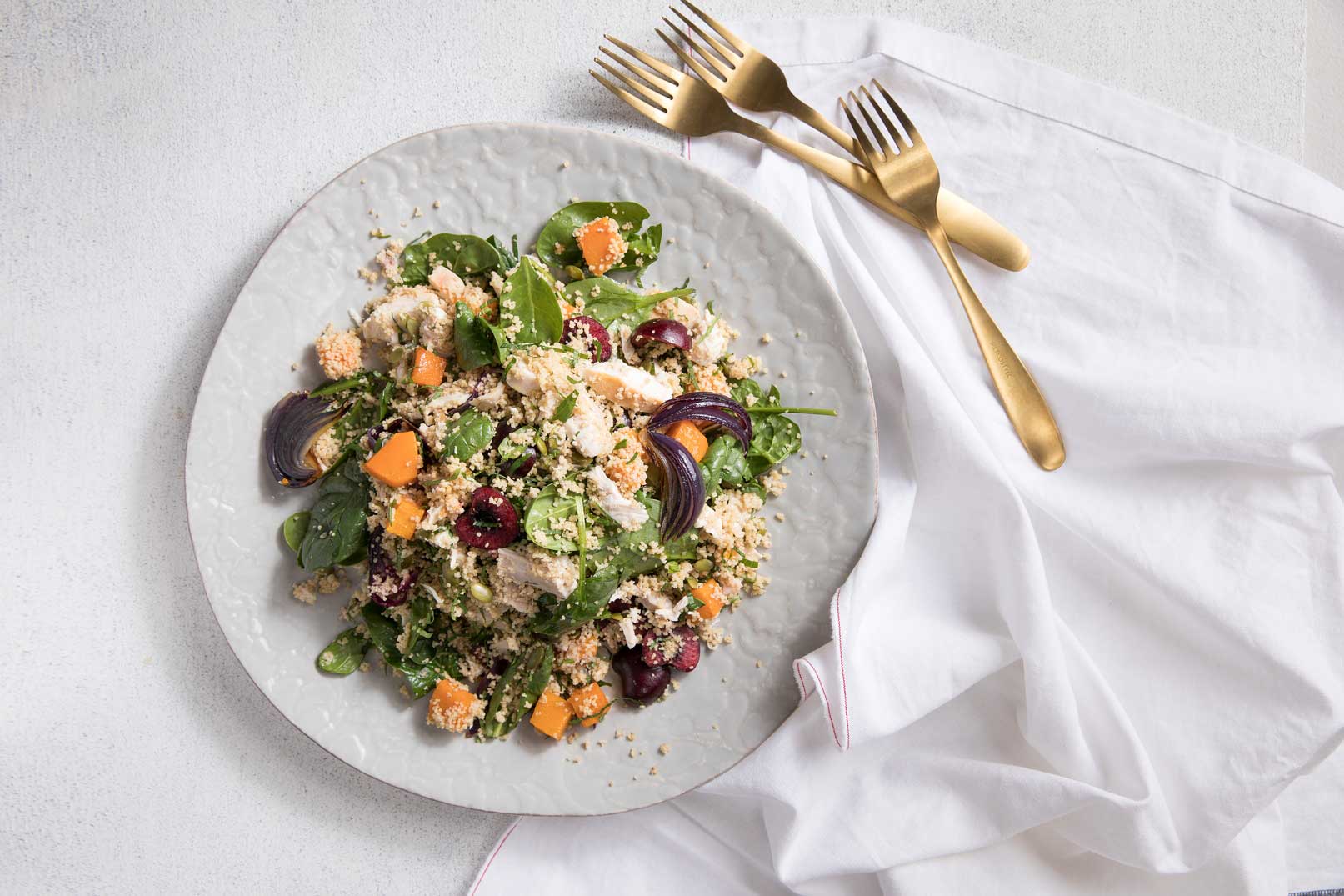 Chicken, cherry and couscous salad on a white plate with 3 gold forks and a white napkin.