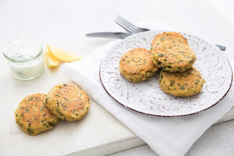 Four couscous cakes on a white plate served on a white napkin with two couscous cakes, dipping sauce and lemon wedges on the side for serving with a knife and fork in the background.