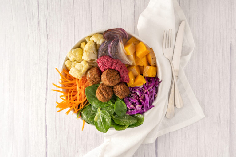 A round white bowl containing falafels, spinach, red cabbage, carrot, cauliflower, red onion, pumpkin with a white linen napkin and a knife and fork on the right.