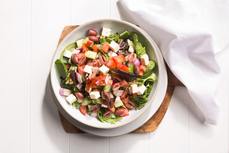 Image of greek salad from above in a white bowl on a wooden chopping board with a white napkin