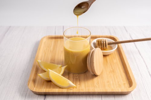 A rectangular timber tray with a glass beaker containing honey mustard dressing with 3 pieces of cut lemon on the left side and a timber honey swizzle on the right.