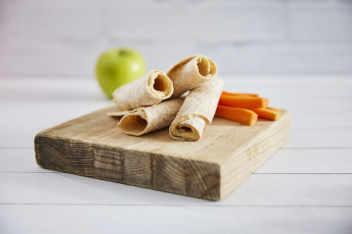 Four pieces of rolled flat bread with tahini inside with carrot sticks on a timber board and a green apple in the background