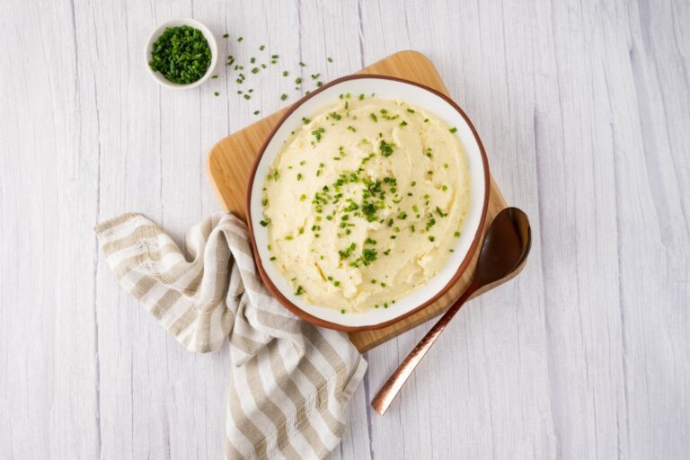 A round shallow bowl containing mashed potato sprinkled with chopped chives sitting on a square timber board. A brown and white striped linen napkin on the left, a serving spoon on the right and a small bowl of chopped chives at the back.