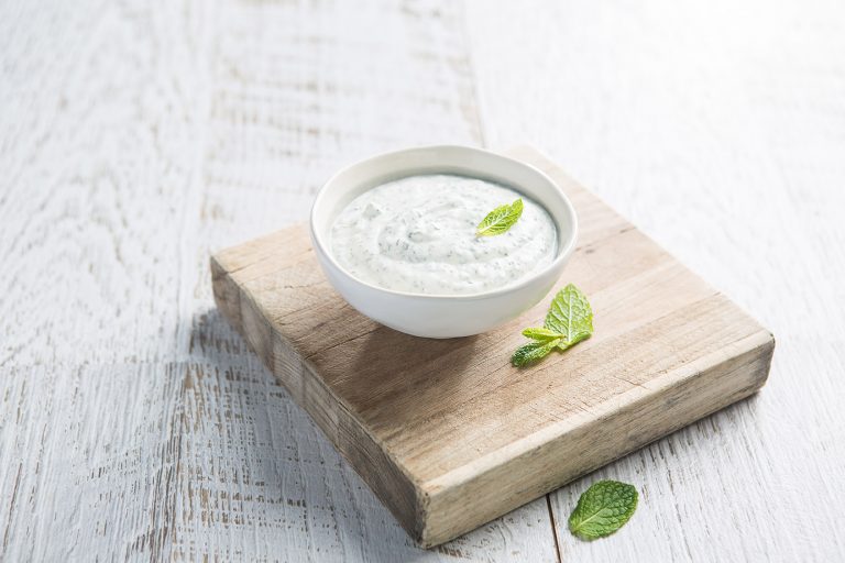 Image of Minty Yoghurt Dip served in a small white bowl on a square cutting board with mint leaves scattered on the side