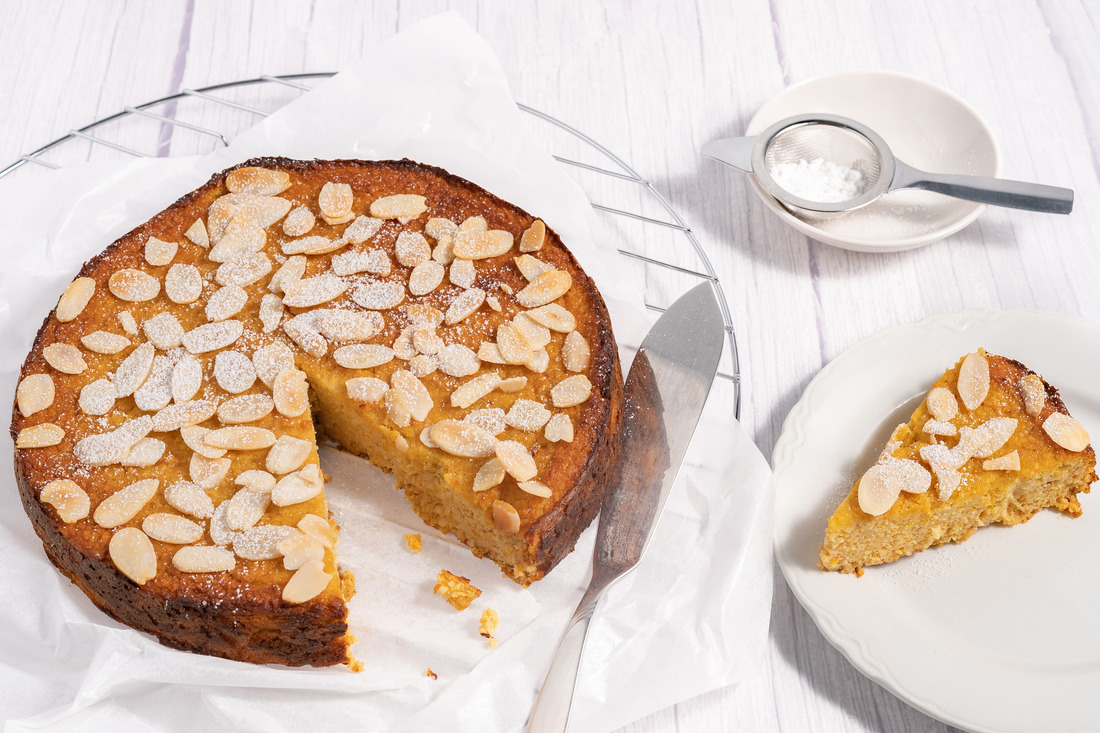 A round orange and almond cake sitting on a white napkin on a round cooling rack. A cut slice of cake on a white saucer to the right.