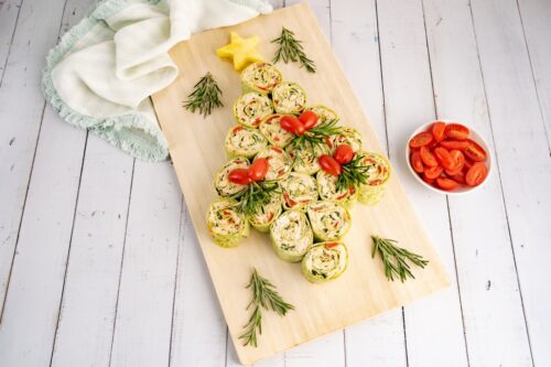 A timber board with pinwheel sandwiches arranged in a Christmas tree shape with a star on the top and garnished with rosemary sprigs. A round white bowl containing halved cherry tomatoes to the right and a fringed fabric napkin on the left.