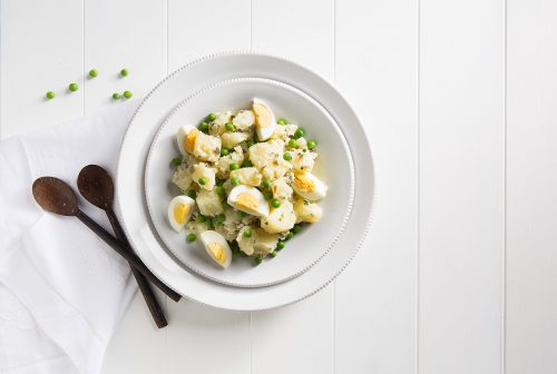 Image of potato salad with egg served in a large white bowl with wooden spoons on the side for serving 