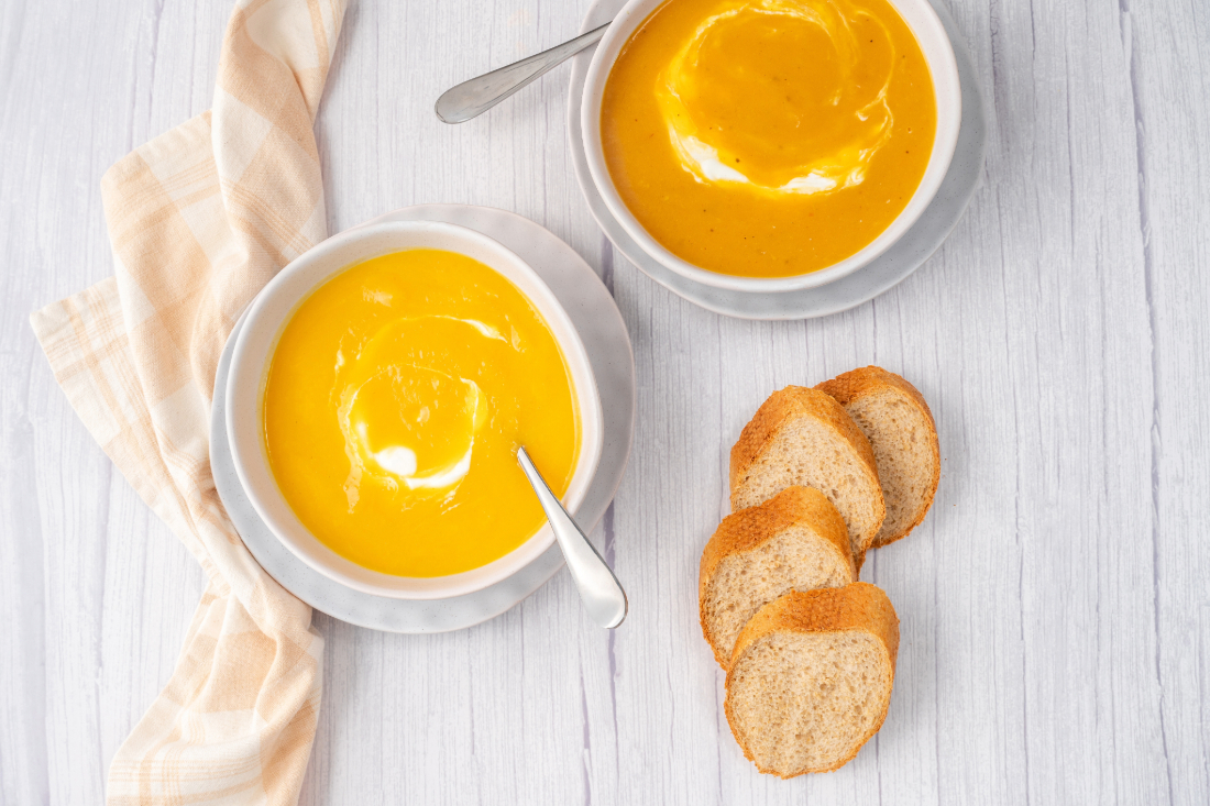 2 round bowls of pumpkin soup with a linen napkin on the left and 4 slices of baguette on the right.