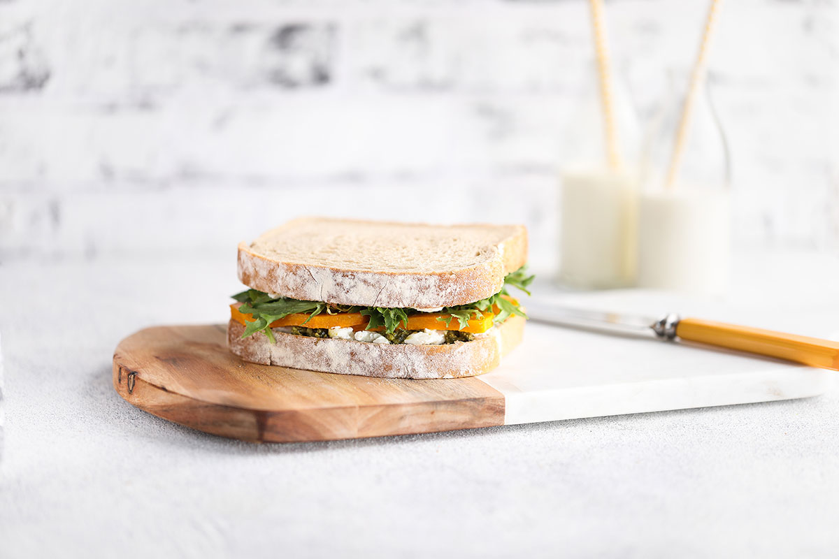 Image of a Pumpkin Cheese Pesto Sandwich served on a wooden and white cutting board with a knife and two bottles of milk with straws in the background