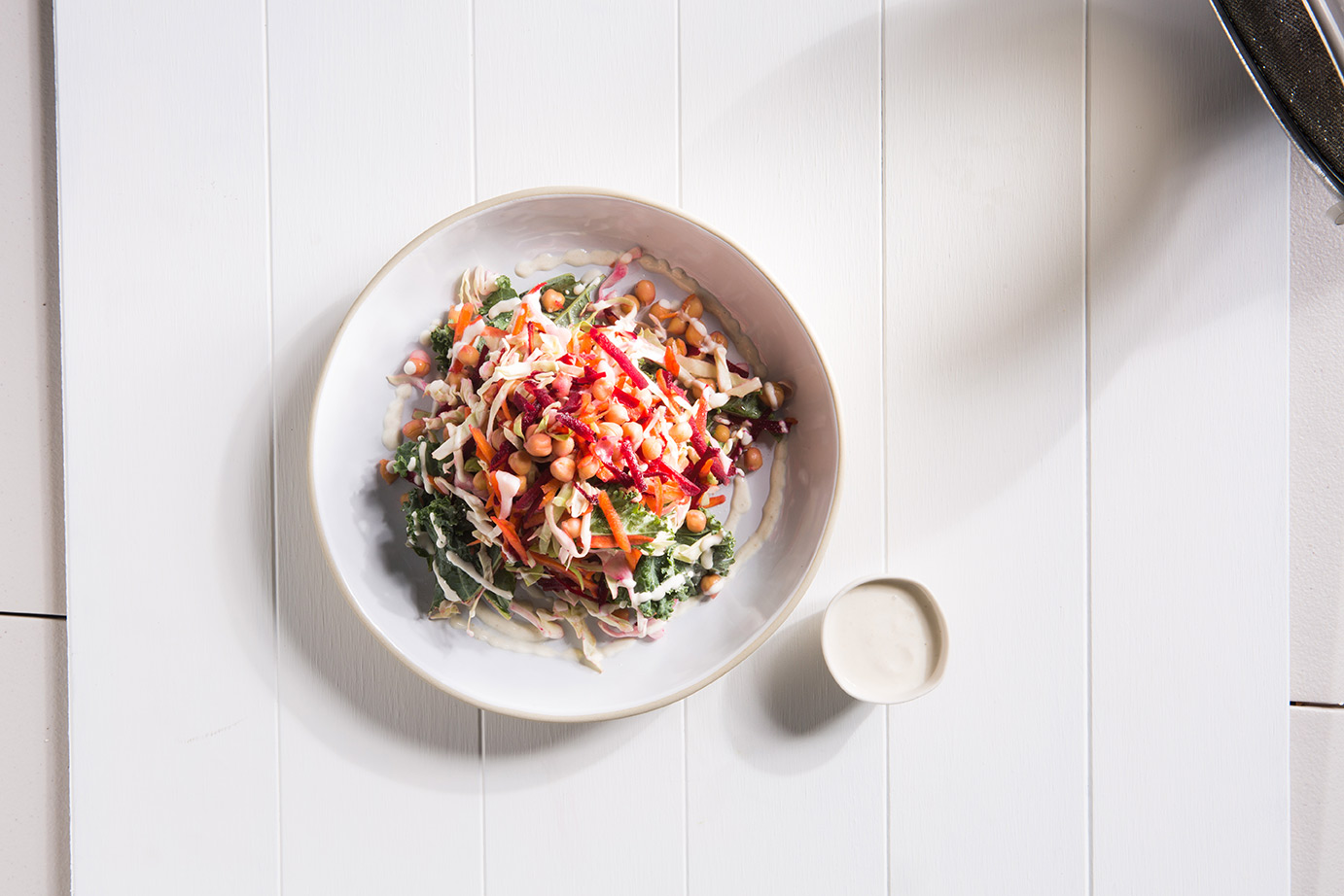 Image of rainbow vegetable salad with chickpeas served in a large white bowl with a small side of dressing, shot from above