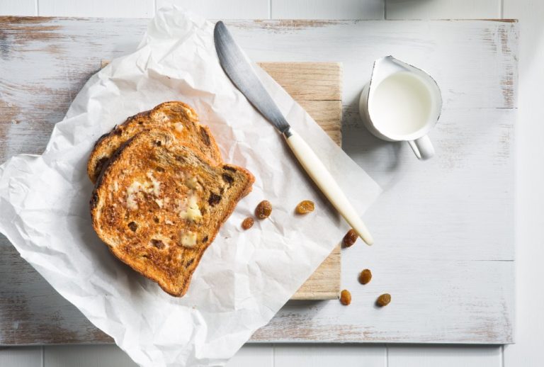 Image two slices of raisin bread on baking paper and a cutting board with a knife and jug of milk on the side