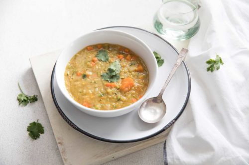 red lentil soup in a white bowl served on a round plate sitting on a white cutting board with a soup spoon, jug of water and white napkin on the side.