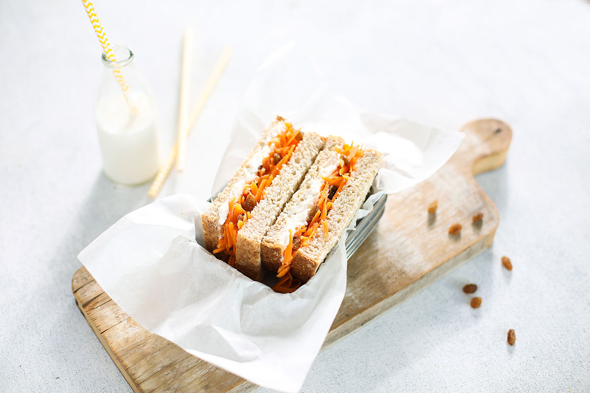 Image of two halves of a ricotta carrot and sultana sandwich served in a metal lunch box with baking paper on a wooden chopping board with a jug of milk and straw in the background