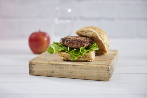 A bread roll containing a meat patty with lettuce on a timber board with a red apple in the background