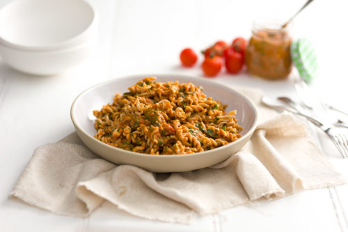 A white bowl containing spiral pasta and sauce with a linen napkin below