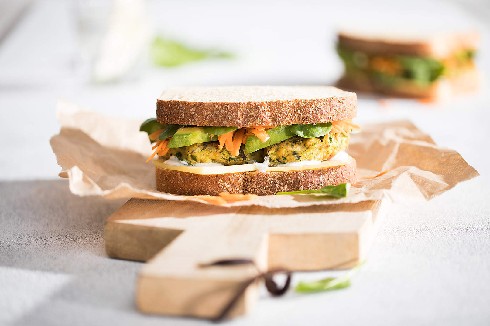 Image of a salmon pattie and avocado sandwich on baking paper served on a wooden cutting board with another sandwich in the background