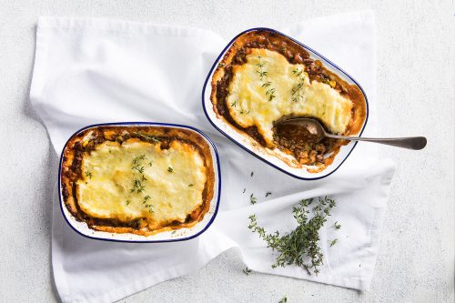 Image of two baked shephard pies sitting on a white cloth napkin and one with a serving spoon, shot from above