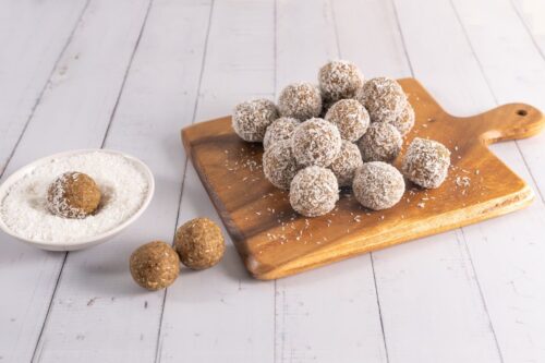 14 round coconut covered bliss balls sitting on a square timber board with a handle. 2 uncoated bliss balls sit to the left with one other bliss ball sitting in a round bowl of desiccated coconut.