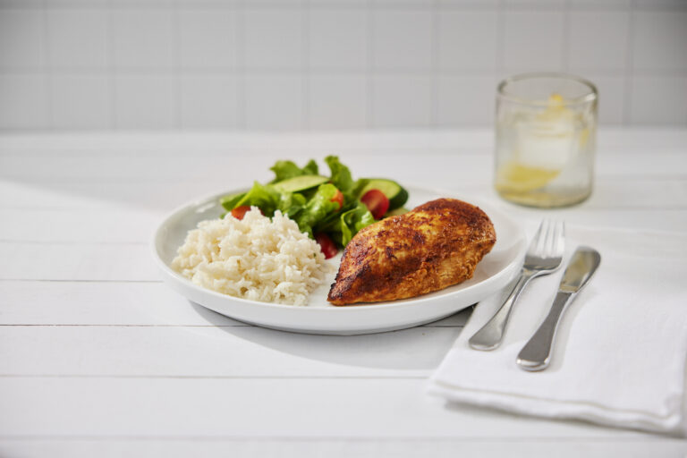 A round white plate with a cooked chicken breast, white rice and salad with a knife and fork on the right