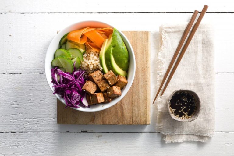 round white bowl containing tofu, brown rice, avocado, cucumber, carrot and red cabbage on a wooden platter with wooden chopsticks and a small bowl of soy sauce on the right