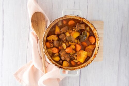 A round casserole dish containing a beef and cannellini bean stew with a wooden spoon and a white napkin to the left.