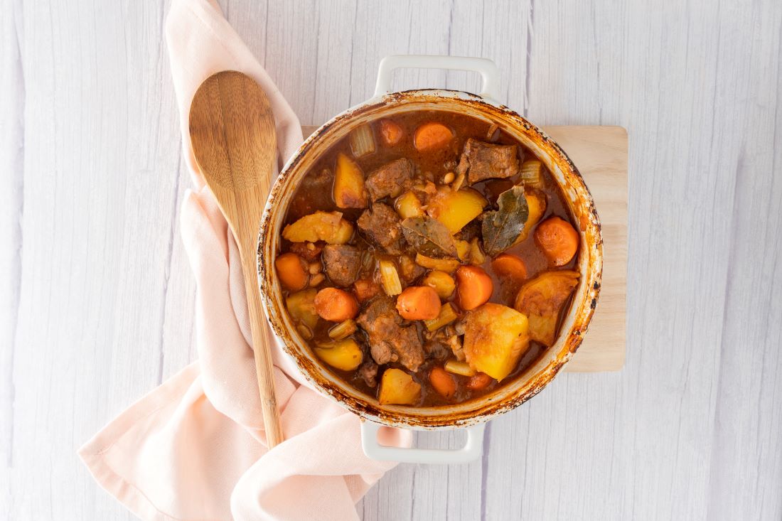 A round casserole dish containing a beef and cannellini bean stew with a wooden spoon and a white napkin to the left.