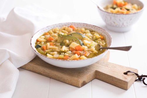 A round white bowl containing vegetable and pasta soup on a timber board