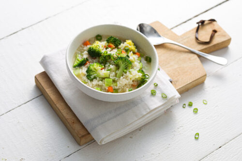 a round white bowl containing rice congee with broccoli served on a wooden platter with a white napkin beneath the bowl and a silver spoon on the right