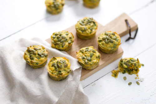 Six vegetable and pepita muffins served on a wooden platter with a white napkin underneath with half a muffin on the right.