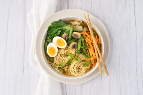 A round white bowl containing ramen noodles, a boiled egg cut in half, sliced mushrooms, shredded carrot and sliced shallots with a pair of timber chopsticks on the right.
