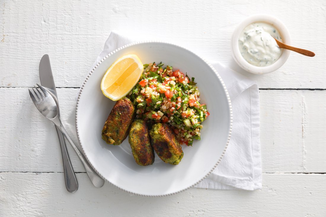 3 vegetable kofta with tabouleh and a quarter of lemon on a round white plate with a small bowl of yoghurt and a white napkin on the right and a silver knife and fork on the left