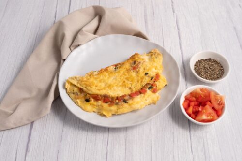 A round white plate with a folded omelette filled with chopped vegetables. To the right a small round bowl containing chopped tomato and another small round bowl containing ground black pepper.