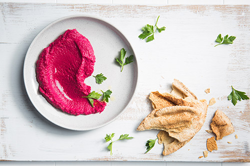 Image of roast beetroot hummus on a serving plate with baked pita on the side and herbs to garnish, shot from above