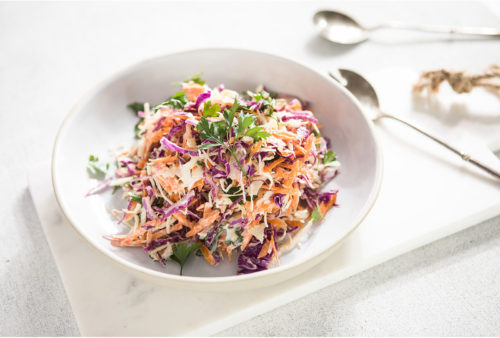 Image of coleslaw in a white bowl served on a white marble cutting board with serving utensils in the background