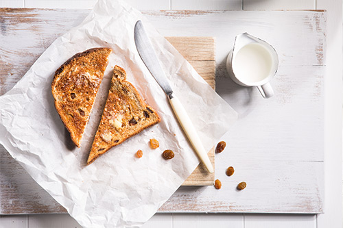 Image one slice of raisin bread sliced into two triangles served on baking paper and a cutting board with a knife and jug of milk on the side